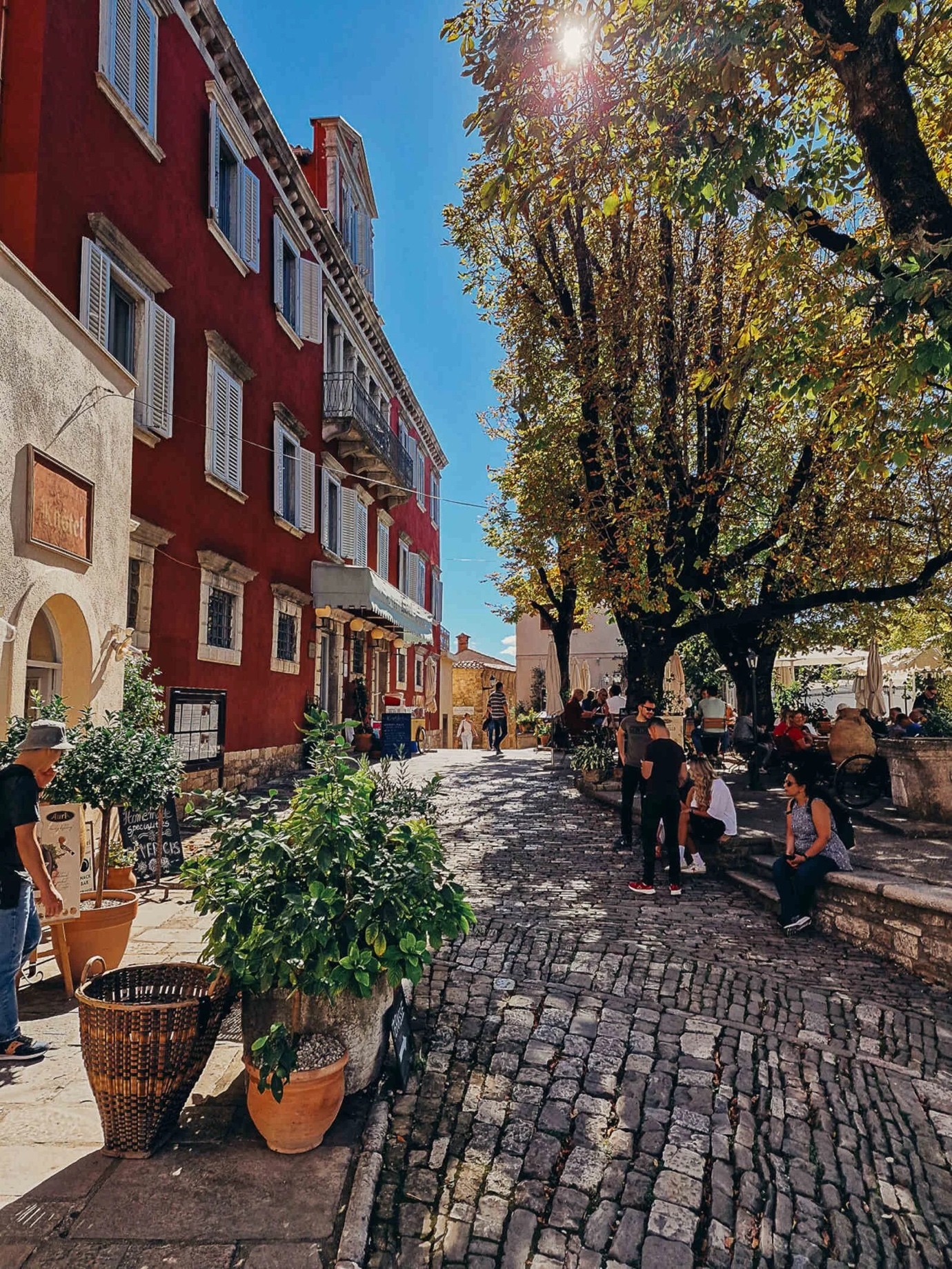 A cobbled street inside the walls of Motovun in autumn, with café tables under a large tree